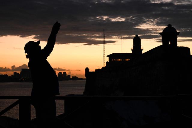 A man takes a photo at Morro Castle at sunset during a blackout in Havana on March 16, 2026. Cuba suffered a widespread power cut on March 16, 2026, according to the national electricity company, against the backdrop of a severe crisis on the island caused by the US energy blockade. (Photo by Yamil LAGE / AFP)