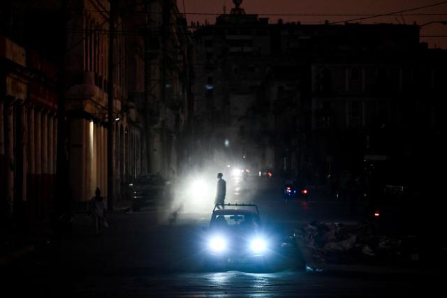 A man walks while cars cruise along a street during a blackout in Havana on March 16, 2026. Cuba suffered a widespread power cut on March 16, 2026, according to the national electricity company, against the backdrop of a severe crisis on the island caused by the US energy blockade. (Photo by Yamil LAGE / AFP)