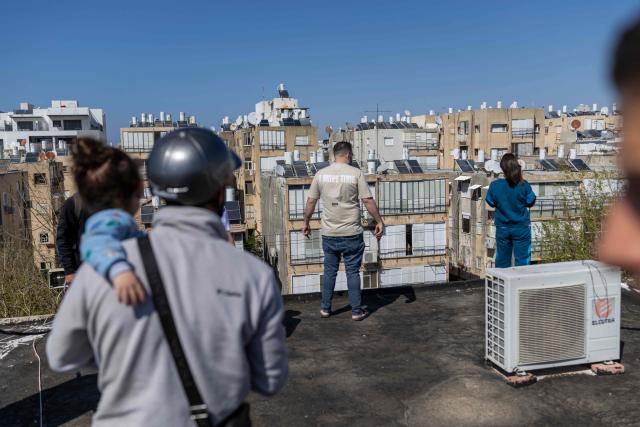People stand on the roof of a house, looking towards the damage following a strike over Holon, on the outskirts of Tel Aviv, on March 17, 2026. On February 28, 2026, Israel and the United States launched strikes on Iran, killing its supreme leader and triggering a war that spread across the Middle East. (Photo by Ilia YEFIMOVICH / AFP) / 