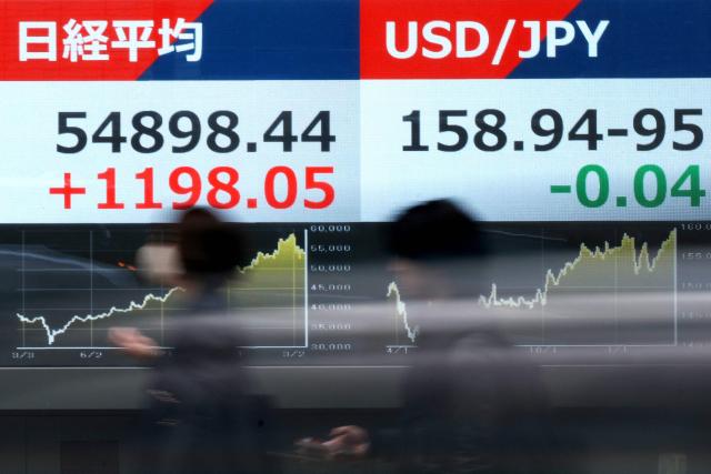 Pedestrians walk past electronic quotation boards displaying the Nikkei Stock Average on the Tokyo Stock Exchange (L) and the foreign exchange rate of the Japanese yen against the US dollar (R) along a street in Tokyo on March 18, 2026. (Photo by Kazuhiro NOGI / AFP)