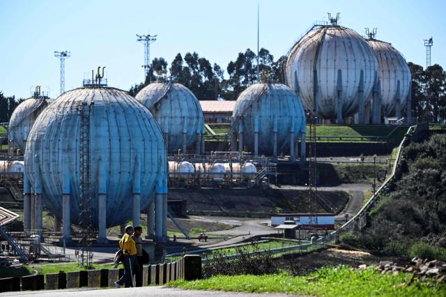 A picture taken on March 17, 2026 shows pedestrians walking next to the Repsol LPG gas plant in the Spanish city of Gijon. The US-Israeli war on Iran has expanded across the Gulf and beyond, upending global energy markets and trade, and virtually halting traffic in the Strait of Hormuz, through which a fifth of the world's crude oil travels. (Photo by ANDER GILLENEA / AFP)