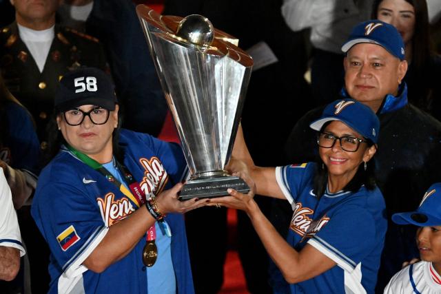 Venezuela’s interim President Delcy Rodriguez (R) and Aracelis Leon, president of the Venezuelan Baseball Federation, hold a trophy during an event to honor the national baseball team after its victory in the World Baseball Classic at the Miraflores Palace in Caracas on March 18, 2026. Venezuelan flags hang from windows, hundreds wear the national team’s jersey, and a holiday has been declared: the country woke up celebrating on March 18 after its first World Baseball Classic title, a celebration that some even link to the fall of Nicolas Maduro. (Photo by Juan BARRETO / AFP)
