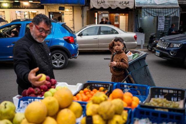 A boy pulling a trash container watches as a man picks out apples from a fruit stand along a street in Aisha Bakkar, a working-class neighbourhood of Beirut, on March 19, 2026. Lebanon was drawn into the Middle East war on March 2 when militant group Hezbollah launched rockets towards Israel to avenge the killing of Iran's supreme leader. Israel responded with intense strikes on Lebanon that have killed at least 968 people and displaced over a million, according to local authorities, and by launching ground operations in the south. (Photo by DIMITAR DILKOFF / AFP)