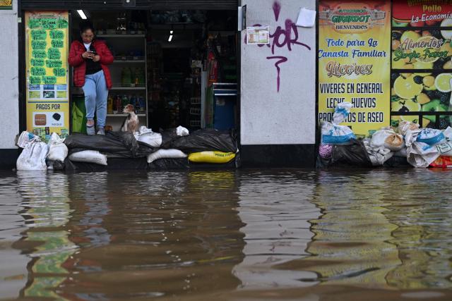 A woman checks her phone at the entrance of a supermarket in front of a flooded street after heavy rains caused rivers to overflow in the Girardot neighborhood of Facatativa municipality, Cundinamarca department, Colombia, on March 19, 2026. (Photo by Raul ARBOLEDA / AFP)