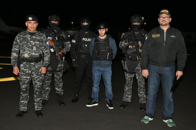 Ecuador’s police officers, alongside Interior Minister John Reimberg (R), pose with Ecuadorian drug trafficker Angel Aguilar (C), alias ‘Lobo Menor’, as he arrives at the Aeropolicial facilities at the airport in Guayaquil on March 19, 2026. Colombian immigration authorities said on March 18, 2026, they had arrested an Ecuadoran drug trafficker linked to the 2023 murder of a popular candidate just before Ecuador's presidential election. (Photo by Marcos PIN / AFP)