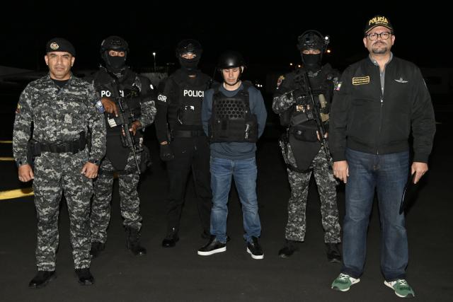 Ecuador’s police officers, alongside Interior Minister John Reimberg (R), pose with Ecuadorian drug trafficker Angel Aguilar (C), alias ‘Lobo Menor’, as he arrives at the Aeropolicial facilities at the airport in Guayaquil on March 19, 2026. Colombian immigration authorities said on March 18, 2026, they had arrested an Ecuadoran drug trafficker linked to the 2023 murder of a popular candidate just before Ecuador's presidential election. (Photo by Marcos PIN / AFP)