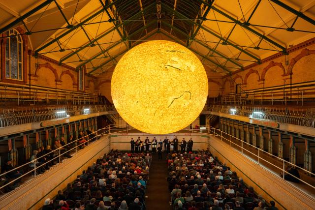 Members of Kantos Chamber Choir perform choral works inspired by the Sun, beneath the artwork entitled 'Helios' by artist Luke Jerram in Victoria Baths, Manchester, north west England on March 19, 2026. The artwork 'Helios' measures seven metres in diameter and has been constructed using approximately 400,000 detailed photographs of the Sun's surface. The piece is a representation of the Sun at an approximate scale of 1:200 million, where each centimetre on the artwork represents 2000km on the Sun's surface. (Photo by Oli SCARFF / AFP)