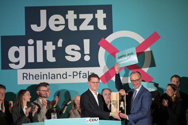CORRECTION / German Chancellor Friedrich Merz (R) is preseneted with wine by an unidentified colleague on stage at the end of a campaign event in Bad Duerkheim, western Germany on March 22, 2026. (Photo by Hannes P. Albert / AFP) / “The erroneous mention[s] appearing in the metadata of this photo by Hannes P. Albert has been modified in AFP systems in the following manner: [unidentified colleague] instead of [Gordon Schnieder]. Please immediately remove the erroneous mention[s] from all your online services and delete it (them) from your servers. If you have been authorized by AFP to distribute it (them) to third parties, please ensure that the same actions are carried out by them. Failure to promptly comply with these instructions will entail liability on your part for any continued or post notification usage. Therefore we thank you very much for all your attention and prompt action. We are sorry for the inconvenience this notification may cause and remain at your disposal for any further information you may require.”