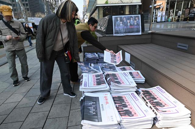 A woman picks up extra edition newspapers reporting the comeback of BTS near the venue of the comeback concert of K-pop boy group BTS in downtown Seoul on March 21, 2026. The world's biggest boy band BTS reunites on March 21, for an open-air comeback concert in the heart of Seoul, expected to draw around 250,000 people and beamed live across the globe. (Photo by Jung Yeon-je / AFP)