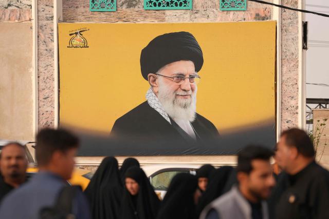 Iraqi Shias walk past a picture of Iran's slain supreme leader Ayatollah Ali Khamenei as they visit the pro-Iranian armed group Kataeb Hezbollah Cemetery in Iraq's central holy city of Najaf as they remember the deceased as well as victims of wars on Eid al-Fitr, marking the end of the fasting month of Ramadan, on March 21, 2026. Iraq has been unwillingly drawn into the regional conflict triggered by the US-Israel attack on its neighbour Iran on February 28. Strikes have targeted Iran-backed groups, which in turn have claimed near-daily attacks on US interests, mostly in Iraq but also across the wider region. (Photo by Qassem al-KAABI / AFP)