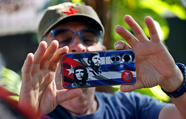 A member of the Nuestra America Convoy takes a photo with his mobile phone, whose case features images of slain Argentine-born Cuban revolutionary Ernesto "Che" Guevara and late Cuban leader Fidel Castro over a Cuban flag, during an event at the Cuban Institute of Friendship with the Peoples (ICAP) in Havana on March 21, 2026. (Photo by Ernesto Mastrascusa / POOL / AFP)