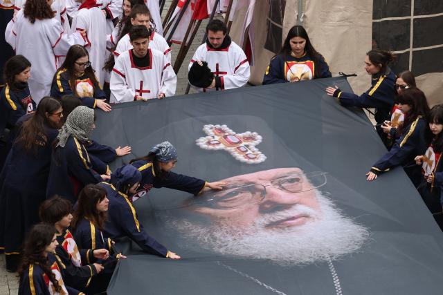 Pilgrims hold a huge banner depicting the late Georgia's Orthodox Patriarch Ilia II during his funeral service outside the Sameba Cathedral in Tbilisi on March 22, 2026. Georgian Orthodox Church leader Ilia II died on March 17, 2026 aged 93, the church said, after nearly half a century at the helm of one of the country's most powerful institutions. (Photo by Giorgi ARJEVANIDZE / POOL / AFP)