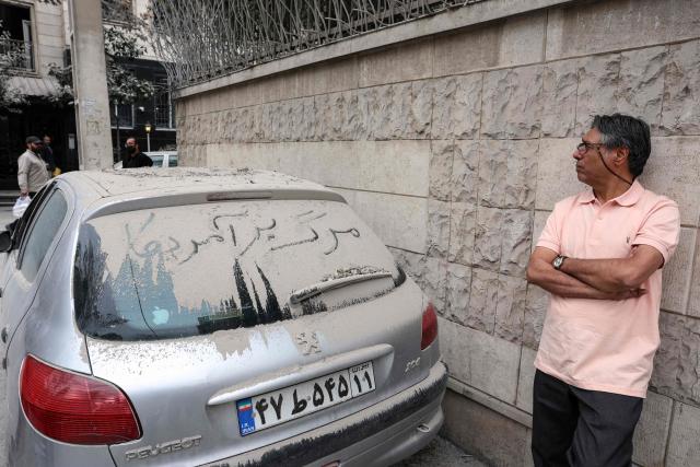 A resident looks at the damage to a destroyed apartment block in northern Tehran as he stands next to a dust-covered car with the words "Down with the USA" written on its rear window on March 23, 2026. A series of strong explosions were heard on March 23, in the Iranian capital, an AFP journalist said, as the war with the United States and Israel raged for its fourth week. On February 28, Israel and the United States launched strikes on Iran killing its supreme leader and triggering a war that spread across the Middle East and unleashed chaos across global markets and sent oil prices soaring. (Photo by AFP) / 