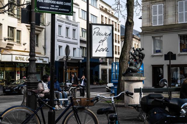 People sit on a bench next to a billboard displaying the title of Canadian singer Celine Dion song "Power of Love" in central Paris on March 23, 2026. "Pour que tu m'aimes encore" or "My Heart Will Go On" can be read on mysterious posters in Paris, also featuring other iconic titles by Celine Dion and suggesting the big return of the Quebec diva for a series of concerts. (Photo by Blanca CRUZ / AFP)