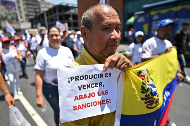 A man holds a sign reading "Producing is winning, down to the snactions!" and  Venezuelan national flag, during a march demanding the complete lifting of US sanctions in Caracas on March 23, 2026. (Photo by Juan BARRETO / AFP)