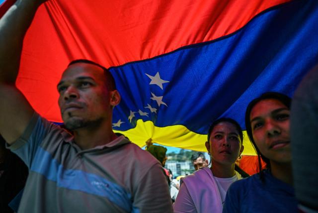 People walk under a huge Venezuelan flag during a march demanding the complete lifting of US sanctions in Caracas on March 23, 2026. (Photo by Juan BARRETO / AFP)