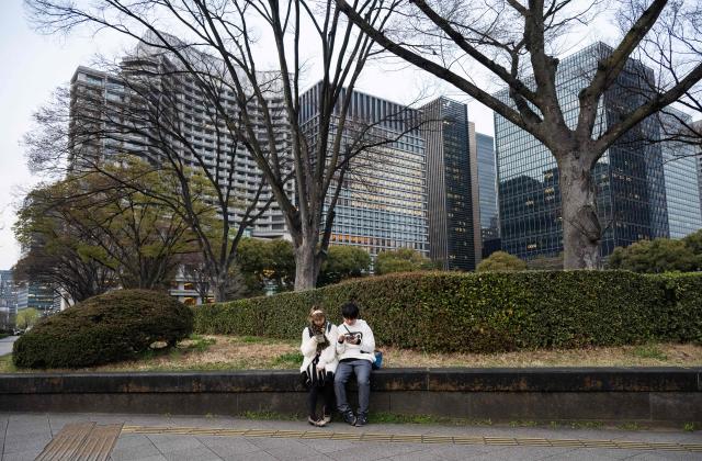 Two people sit on a wall looking at their phones at the Kokyo Gaien National Garden in Tokyo on March 24, 2026. (Photo by Andrew CABALLERO-REYNOLDS / AFP)