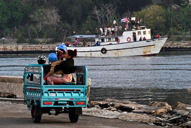 People ride an electric three wheel motorcycle as the vessel Maguro, arriving from Mexico with humanitarian aid as part of the Nuestra America convoy, docks at the port of Havana on March 24, 2026. (Photo by YAMIL LAGE / AFP)