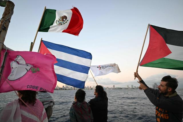 Activists wave Cuban, Mexican and Palestinian flags on board the vessel Maguro as it arrives from Mexico with humanitarian aid as part of the Nuestra America convoy, docking at the port of Havana on March 24, 2026. The first boat of a flotilla carrying medical supplies, food and solar panels reached Cuba on March 24, 2026 to aid the island as a US fuel blockade deepens its energy crisis. The Maguro shrimp fishing boat docked in Havana three days later than hoped after battling strong winds, currents and a pesky battery, with two other ships due to follow. (Photo by YURI CORTEZ / AFP)