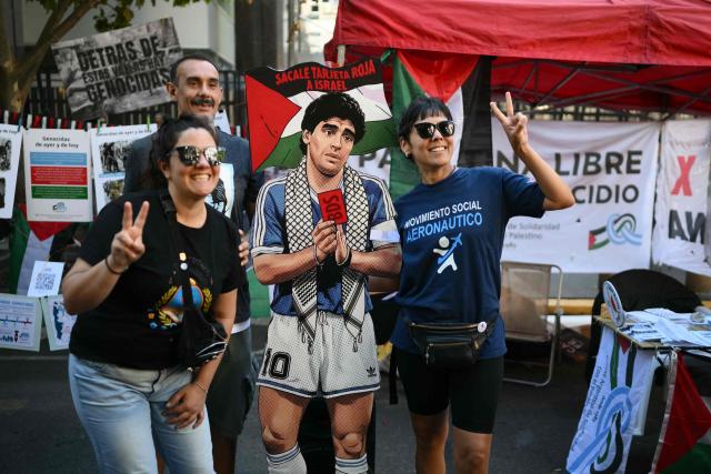 People flash the V sign as they pose with a cutout depicting Argentine football legend Diego Maradona holding a red card and a sign reading “Give Israel a red card” during a demonstration in front of the Israeli Embassy in Buenos Aires on March 24, 2026, as part of the mobilization marking the 50th anniversary of the military coup. In Argentina, March 24th is a day of mourning, marches and political disputes. Fifty years on from the coup d’etat, thousands of people are taking to the streets again to commemorate the victims of a dictatorship that the government of far-right leader Javier Milei is seeking to rewrite. (Photo by Luis ROBAYO / AFP)
