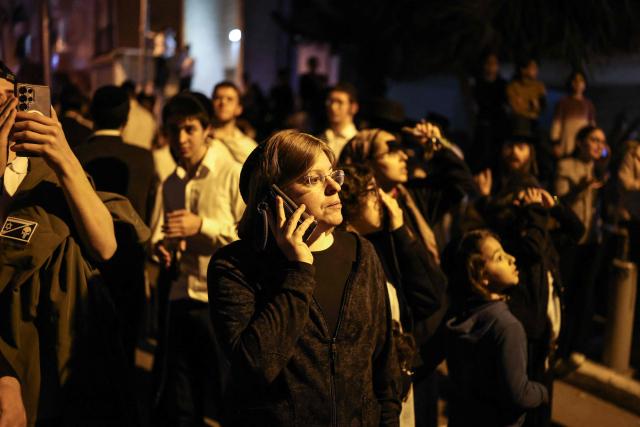 A woman uses a mobile phone as she stands at the site where a building was struck by a projectile in Bnei Brak, on the eastern outskirts of Tel Aviv, on March 24, 2026. The Middle East was sparked by joint US-Israeli strikes on Iran that triggered a wave of retaliatory missile and drone attacks against Israel and several other countries in the region. (Photo by Ilia YEFIMOVICH / AFP) / 