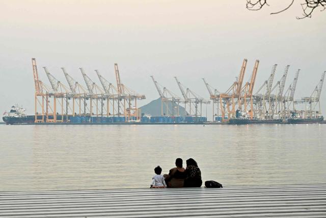 (FILES) A family sits against the backdrop of a dockyard off coast city of Fujairah, in the Strait of Hormuz in the northern Emirate on February 25, 2026. The US president sent a peace plan to Iran as he voiced optimism on March 25, 2026 at ending nearly a month of warfare, with Tehran announcing that it will let "non-hostile" oil vessels go through the crucial Strait of Hormuz. Oil prices dropped sharply and stocks in Asia rose on broader de-escalation hopes following nearly four weeks of war, as the US president appeared to be ramping up efforts to bring an end to his joint military operation with Israel. (Photo by Giuseppe CACACE / AFP)