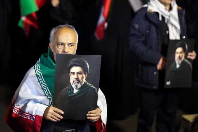 A man draped in a national flag holds a portrait of Iran's supreme leader Mojtaba Khamenei as people march in support of the Iranian armed forces in central Tehran on March 25, 2026. President Donald Trump is ready to "unleash hell" if Iran doesn't accept a deal to end the Middle East war, the White House warned on March 25, adding that talks continued despite Tehran reportedly rejecting a proposed US peace plan. (Photo by AFP) / 
