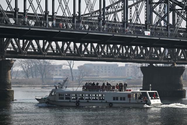 A Chinese tour boat cruises on the Yalu River past the Sino-Korean Friendship Bridge and the Yalu River Broken Bridge as the North Korean town of Sinuiju is seen behind, opposite the border city of Dandong, in China's northeast Liaoning province on March 26, 2026. Passenger rail travel between China and North Korea resumed on March 12, after a pause of more than six years triggered by the COVID-19 outbreak. (Photo by GREG BAKER / AFP)