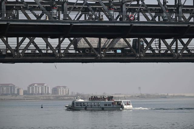A Chinese tour boat cruises on the Yalu River below a truck on the Sino-Korean Friendship Bridge as the North Korean town of Sinuiju is seen behind, opposite the border city of Dandong, in China's northeast Liaoning province on March 26, 2026. Passenger rail travel between China and North Korea resumed on March 12, after a pause of more than six years triggered by the COVID-19 outbreak. (Photo by GREG BAKER / AFP)