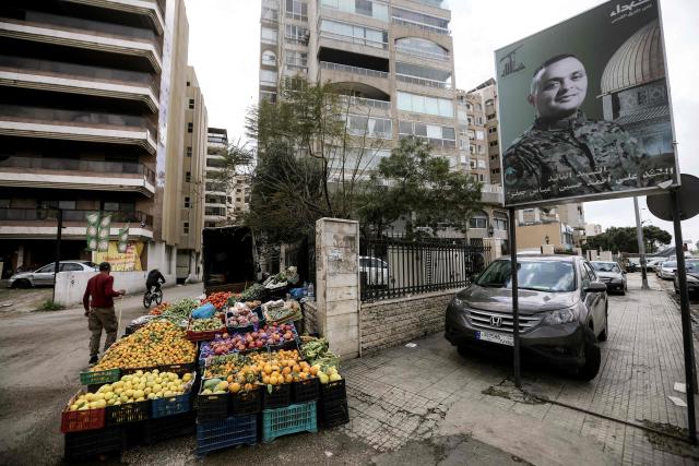 A street vendor stands by his stall next to a billboard bearing the portrait of Ali Ahmed Hussein, a Hezbollah commander who was killed in southern Lebanon 2024, in the Shayyah neighbourhood of Beirut's southern suburbs on March 27, 2026. Lebanon was drawn into the Middle East war on March 2 when Tehran-backed Hezbollah began firing rockets into Israel to avenge the killing of the Iranian supreme leader. Israel has since been bombing Lebanon, mainly in areas where Hezbollah has long held sway, and has sent in ground troops in a push to establish a buffer zone in south Lebanon. (Photo by ibrahim AMRO / AFP) / 
