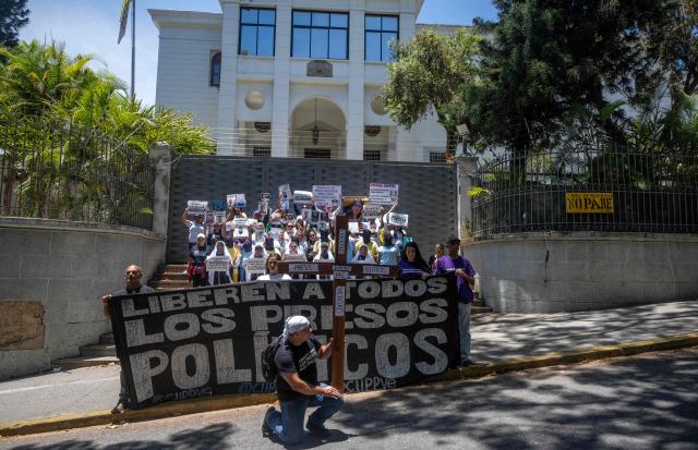 A relative of a political prisoner carries a large wooden cross with words such as "Freedom," "Justice," "Peace," "Truth," and "Political Prisoners" during a march in Caracas on March 27, 2026, as a banner reading "Free all political prisoners" is held in the background. Dressed in black and white robes, mothers of political prisoners marched on Friday in Caracas to the headquarters of the nunciature to ask the Vatican to intercede for an amnesty for their relatives. (Photo by Maryorin Mendez / AFP)