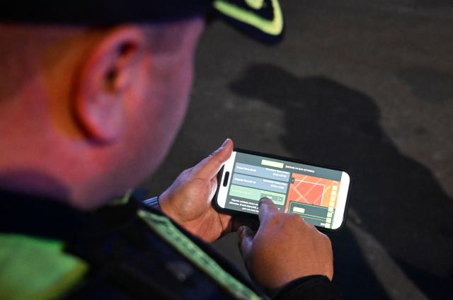 A Colombian police officer uses an app on his mobile phone as he patrols the San Bernardo neighborhood, a critical, high-risk zone known for micro-trafficking, insecurity, and social conflict in downtown Bogota on March 26, 2026. Colombian police use an artificial intelligence tool to predict crime hotspots in real time and create security plans based on large amounts of geographic data. The software, called "IAPol," shows on a map the areas where different risk factors come together, like crime statistics, emergency calls, and police presence, allowing for quicker planning in a country with high crime rates. (Photo by Raul ARBOLEDA / AFP)