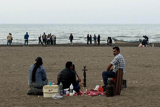 A man smokes a waterpipe (shisha or hookah) while sitting with others along a beach by the Caspian Sea coast in Sorkhrud in Iran's northern Mazandaran province on March 24, 2026, where many families have relocated due to the ongoing war with the US and Israel. Israel and the United States launched strikes on Iran on February 28, killing the Islamic republic's supreme leader and sparking a war that has since spread across the Middle East. (Photo by ATTA KENARE / AFP) / 
