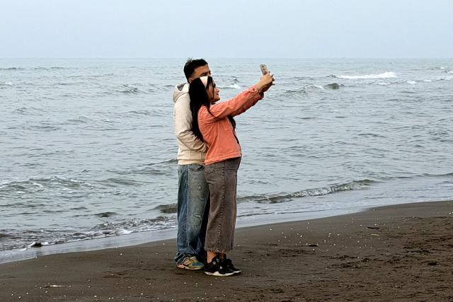 A couple poses for a "selfie" photo on a phone along a beach by the Caspian Sea coast in Sorkhrud in Iran's northern Mazandaran province on March 24, 2026, where many families have relocated due to the ongoing war with the US and Israel. Israel and the United States launched strikes on Iran on February 28, killing the Islamic republic's supreme leader and sparking a war that has since spread across the Middle East. (Photo by ATTA KENARE / AFP) / 