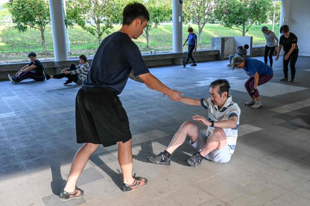 This photograph taken on March 17, 2026 shows coach Tan Shie Boon (L) warming up with Singaporean elders before a parkour training session in Singapore. This is "geriatric parkour", where around 20 retirees learned to tackle a series of relatively demanding exercises, building their agility and enjoying a sense of camaraderie. (Photo by Roslan RAHMAN / AFP) / To go with "Singapore-Ageism" Focus by Martin ABBUGAO