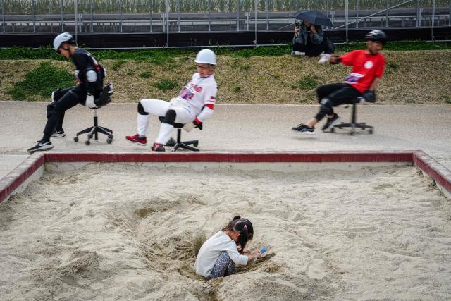 A child (front) plays in the sandbox as racers riding on office chairs compete in the ISU-1 Grand Prix in Kyotanabe, Kyoto prefecture on March 29, 2026. The ISU-1 Grand Prix is an endurance race supervised by the Japan Office Chair Racing Association (JORA), in which participants compete to see how many laps they can complete on office chairs over two hours. The event originated in 2010 in the Kirara shopping district of Kyotanabe, Kyoto Prefecture, and is now held at more than 20 locations across Japan. (Photo by Yuichi YAMAZAKI / AFP)