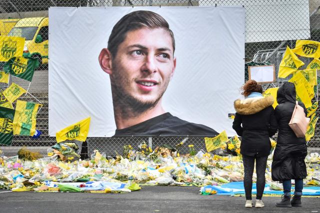 (FILES) People look at yellow flowers displayed in front of the portrait of Argentinian forward Emiliano Sala at the Beauvoir stadium in Nantes, on February 8, 2019. The commercial court of Nantes is due to deliver its decision on March 30, 2026 regarding the financial dispute between the two clubs, seven years after the death of Emiliano Sala in a plane crash. Sala, 28, died in a plane crash having inhaled toxic levels of carbon monoxide from the aircraft's faulty exhaust system during an unlicensed 2019 flight en route from Nantes to Cardiff. (Photo by LOIC VENANCE / AFP)