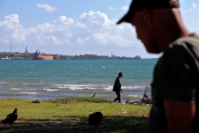 Men walk along the shore of the Bay of Matanzas as tanker ships are seen in the background at the Supertanker Base in the Industrial Zone of the port of Matanzas, Cuba, on March 30, 2026. US President Donald Trump said on March 29, 2026, that Moscow could send oil to Cuba despite Washington's de facto fuel blockade, as a Russian tanker was expected to deliver some much-needed crude to the crisis-hit island. The Anatoly Kolodkin, which is carrying 730,000 barrels of crude, was off northeast Cuba on the evening of March 29 and is expected to dock in the western port of Matanzas by March 31, according to shipping tracker MarineTraffic. (Photo by AFP)
