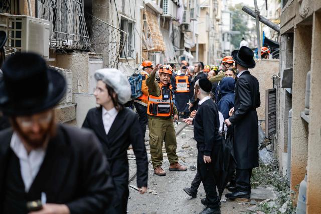 A first responder gestures as Ultra-Orthodox Jews stand at the scene of an impact following an Iranian strike over Bnei Brak in central Israel on April 1, 2026. The Israeli military said its air defences responded to a missile attack from Iran on April 1, with warning sirens activated across central Israel. (Photo by Marco Longari / AFP) / 
