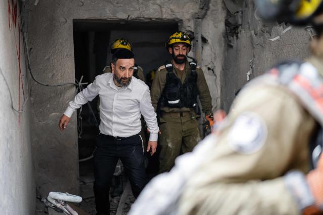 A man walking alongside first responders surveys the damage at the scene of an impact following an Iranian strike over Bnei Brak in central Israel on April 1, 2026. The Israeli military said its air defences responded to a missile attack from Iran on April 1, with warning sirens activated across central Israel. (Photo by Marco Longari / AFP) / 