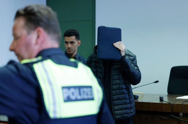 Defendant 71-year-old Joachim K (R) hides his face as he is brought to the courtroom prior to the start of a trial against him and two others for backing the self-styled "United Patriots" group, part of Germany's extremist Reichsbuerger scene, on April 1, 2026 at the higher Regional court of Munich, southern Germany. The group, referred to by investigators as the Kaiserreichsgruppe or "Imperial Group", was allegedly committed to forcibly establishing a new authoritarian Germany based on the old 1871 Imperial Constitution. (Photo by Michaela Stache / AFP) / GERMAN COURT REQUESTS THAT THE FACE OF THE DEFENDANT MUST BE MADE UNRECOGNISABLE