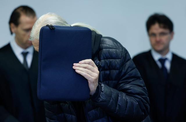 Defendant 71-year-old Joachim K (C) hides his face as he arrives at the courtroom prior to the start of a trial against him and two others for backing the self-styled "United Patriots" group, part of Germany's extremist Reichsbuerger scene, on April 1, 2026 at the higher Regional court of Munich, southern Germany. The group, referred to by investigators as the Kaiserreichsgruppe or "Imperial Group", was allegedly committed to forcibly establishing a new authoritarian Germany based on the old 1871 Imperial Constitution. (Photo by Michaela Stache / AFP) / GERMAN COURT REQUESTS THAT THE FACE OF THE DEFENDANT MUST BE MADE UNRECOGNISABLE