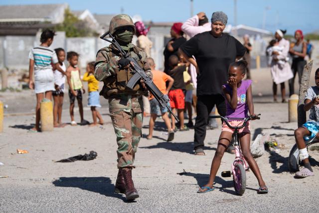 A soldier of the South African National Defence Force (SANDF) walks past local residents during a patrol as part of Operation Prosper in Mitchells Plain, near Cape Town, on April 1, 2026. Operation Prosper sees the SANDF play a supporting role to the South African Police Service (SAPS) in crime prevention operations. (Photo by RODGER BOSCH / AFP)