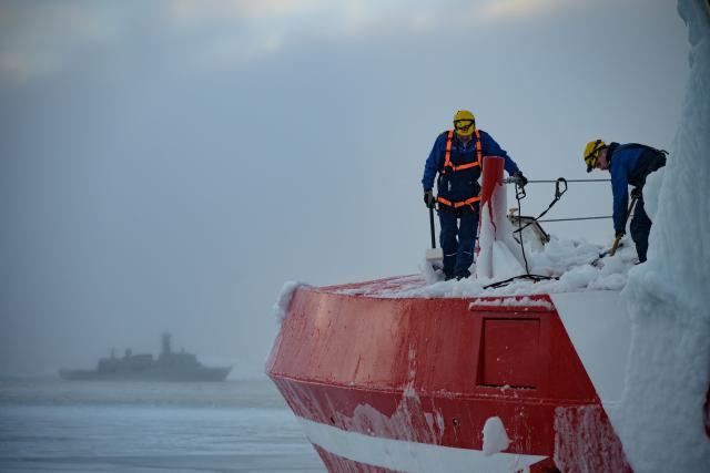 Crew members of the Sarfaq Ittuk ferry de-ice the bow of the vessel after it became stuck in the ice, while a Danish frigate fades into the mist in the background, in Sisimiut, Greenland, on March 15, 2026. The Sarfaq Ittuk is a vital and iconic coastal ferry for Greenlanders, keeping small west coast communities connected to low cost. It links Qaqortoq in the south to Ilulissat in the north in a week-long journey. This year, an unusually warm January allowed the ferry to reach Ilulissat much earlier than usual, as the town is normally blocked by sea ice in March. Around 200 passengers travel on board for medical needs, work, tourism, or simply to meet people. But with new airports opening and a lack of investors, the future of the Sarfaq Ittuk is increasingly uncertain. (Photo by Florent VERGNES / AFP)