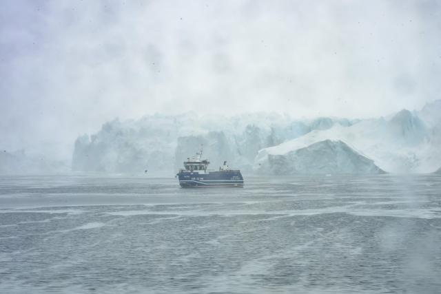 This photograph, taken through a porthole, shows a fishing boat sailing past icebergs that have drifted out of Ilulissats Icefjord, Greenland, on March 15, 2026. The Sarfaq Ittuk is a vital and iconic coastal ferry for Greenlanders, keeping small west coast communities connected to low cost. It links Qaqortoq in the south to Ilulissat in the north in a week-long journey. This year, an unusually warm January allowed the ferry to reach Ilulissat much earlier than usual, as the town is normally blocked by sea ice in March. Around 200 passengers travel on board for medical needs, work, tourism, or simply to meet people. But with new airports opening and a lack of investors, the future of the Sarfaq Ittuk is increasingly uncertain. (Photo by Florent VERGNES / AFP)