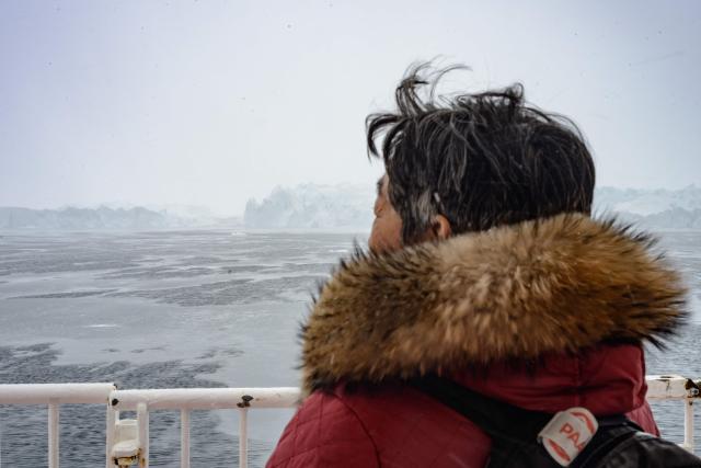 A woman looks out from the deck of the Sarfaq Ittuk ferry at the icebergs of the Icefjord, near Ilulissat, Greenland, on March 15, 2026. The Sarfaq Ittuk is a vital and iconic coastal ferry for Greenlanders, keeping small west coast communities connected to low cost. It links Qaqortoq in the south to Ilulissat in the north in a week-long journey. This year, an unusually warm January allowed the ferry to reach Ilulissat much earlier than usual, as the town is normally blocked by sea ice in March. Around 200 passengers travel on board for medical needs, work, tourism, or simply to meet people. But with new airports opening and a lack of investors, the future of the Sarfaq Ittuk is increasingly uncertain. (Photo by Florent VERGNES / AFP)
