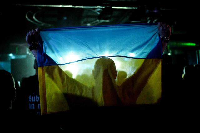 A spectator holds a Ukrainian national flag during a concert of the Ukrainian metal band "1914" performing on stage during a concert at the Proxima music club in Warsaw on March 25, 2026. Like other bands, "1914" is raising money for the Ukrainian army. Kumar is attempting to "open the eyes" of Europeans in light of the Kremlin's warmongering, showing that Ukraine poses as an "eastern shield". Their latest album, released in November 2025, follows the story of a Ukrainian from Galicia in their fight against Russia in the First World War. (Photo by Sergei GAPON / AFP)