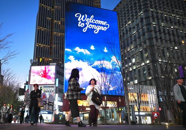 People walk in front of digital billboards at Gwanghwamun Square in Seoul on April 1, 2026. Hopes of making downtown Seoul dazzle more than Times Square have hit a setback with new guidelines to dim the massive digital billboards that light up the South Korean capital after a barrage of complaints. (Photo by Jung Yeon-je / AFP)
