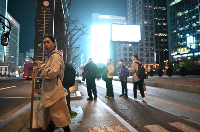 People wait at a bus stop in front of digital billboards near Gwanghwamun Square in Seoul on April 1, 2026. Hopes of making downtown Seoul dazzle more than Times Square have hit a setback with new guidelines to dim the massive digital billboards that light up the South Korean capital after a barrage of complaints. (Photo by Jung Yeon-je / AFP)