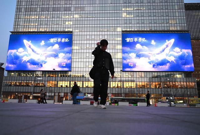 People walk in front of digital billboards at Gwanghwamun Square in Seoul on April 1, 2026. Hopes of making downtown Seoul dazzle more than Times Square have hit a setback with new guidelines to dim the massive digital billboards that light up the South Korean capital after a barrage of complaints. (Photo by Jung Yeon-je / AFP)